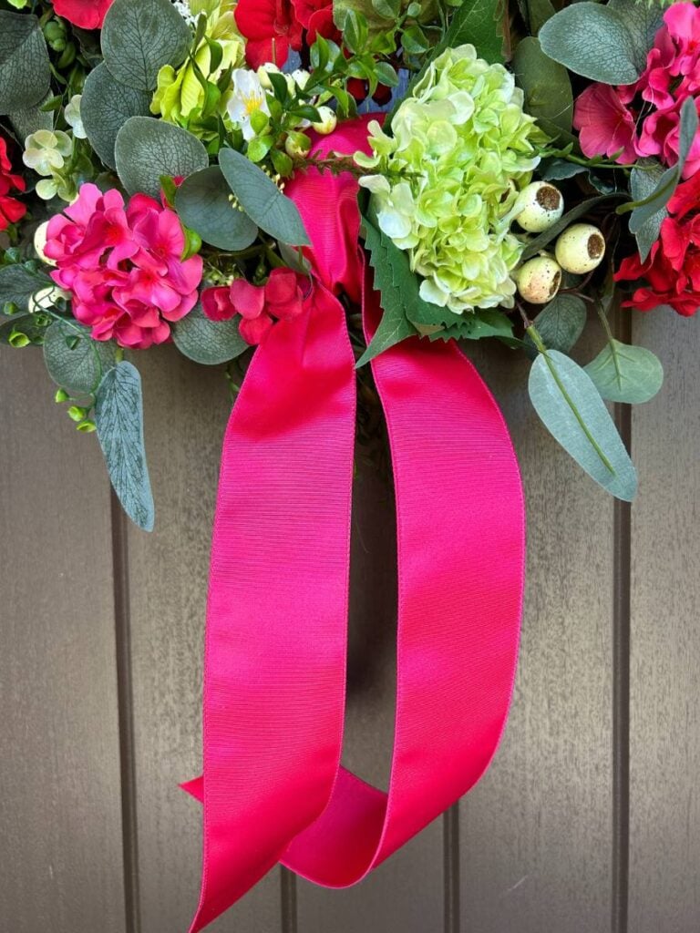 fuchsia satin ribbon tied in a knot on a grapevine wreath covered with red, deep pink, and lime green spring and summer blooms