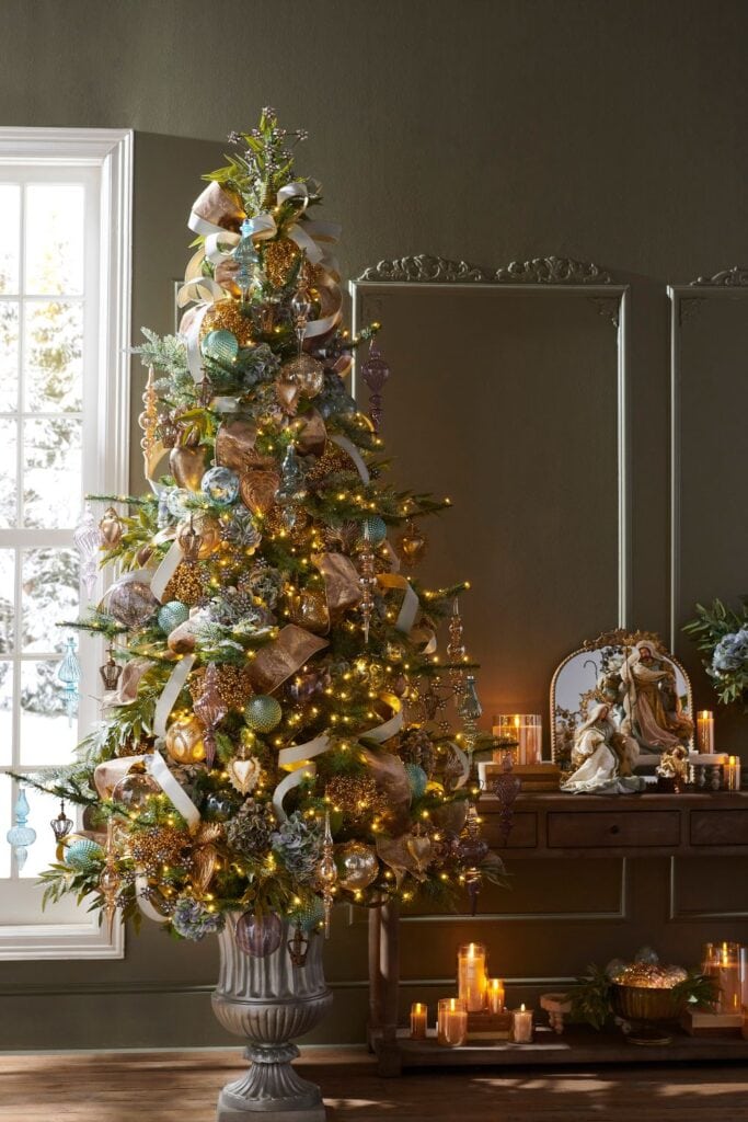 The "Oh Night Divine" tree is elegant and rich in style. It’s decorated with gold, bronze, and soft blue ornaments, along with large ribbons. The tree has warm lights that highlight the beautiful metallic ornaments. It stands in a tall urn, adding a grand touch, and is surrounded by candles and a nativity scene for a peaceful, classic Christmas look.