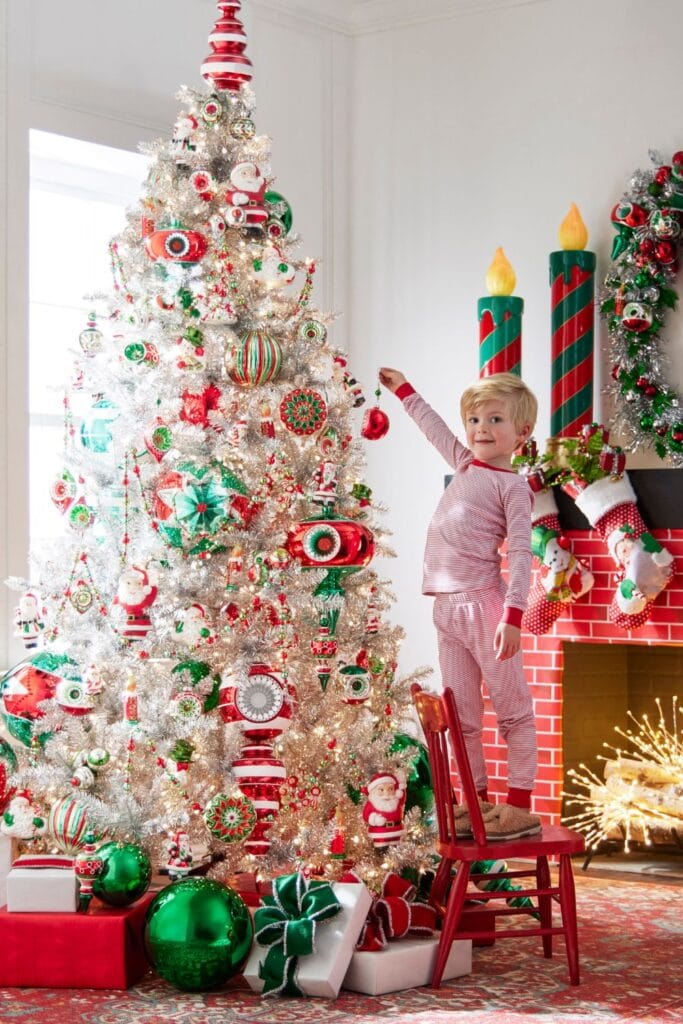 The "Tinsel Town" tree is bright and festive, with red, green, and white decorations, shiny tinsel, and playful ornaments. Surrounding the tree are matching holiday accents like a Santa wreath, oversized candles, and stockings, creating a lively, cheerful Christmas atmosphere.