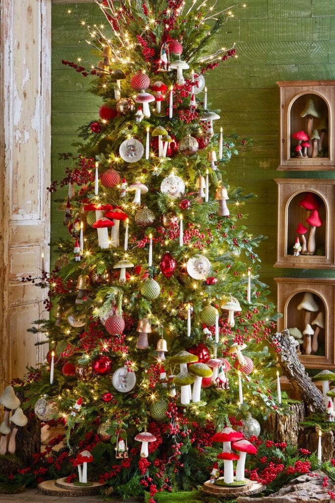 The "Woodland Wonder" tree is decorated with nature-inspired elements. It features red and green ornaments, berries, and candles, along with charming mushroom decorations throughout. The tree has a natural, woodsy feel, with details that evoke a forest setting. The surrounding space includes small mushroom displays and logs, adding to the rustic, woodland theme. The warm lights and natural colors create a cozy, festive atmosphere.