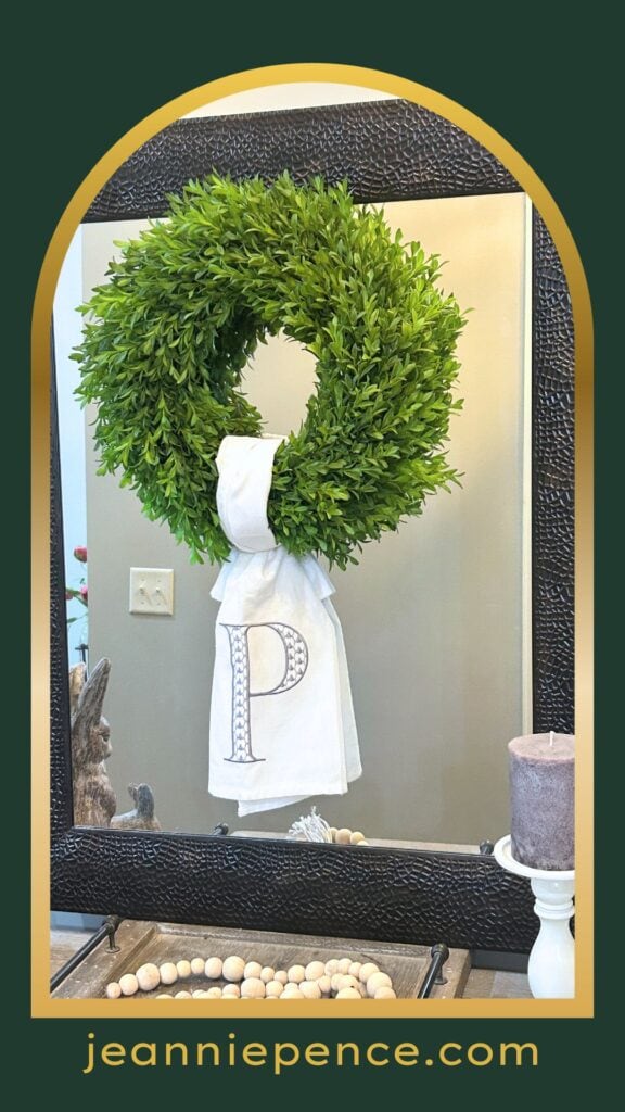 green boxwood wreath with a monogrammed tea towel used as a wreath sash, hanging in front of a rectangular mirror with hammered metal frame
