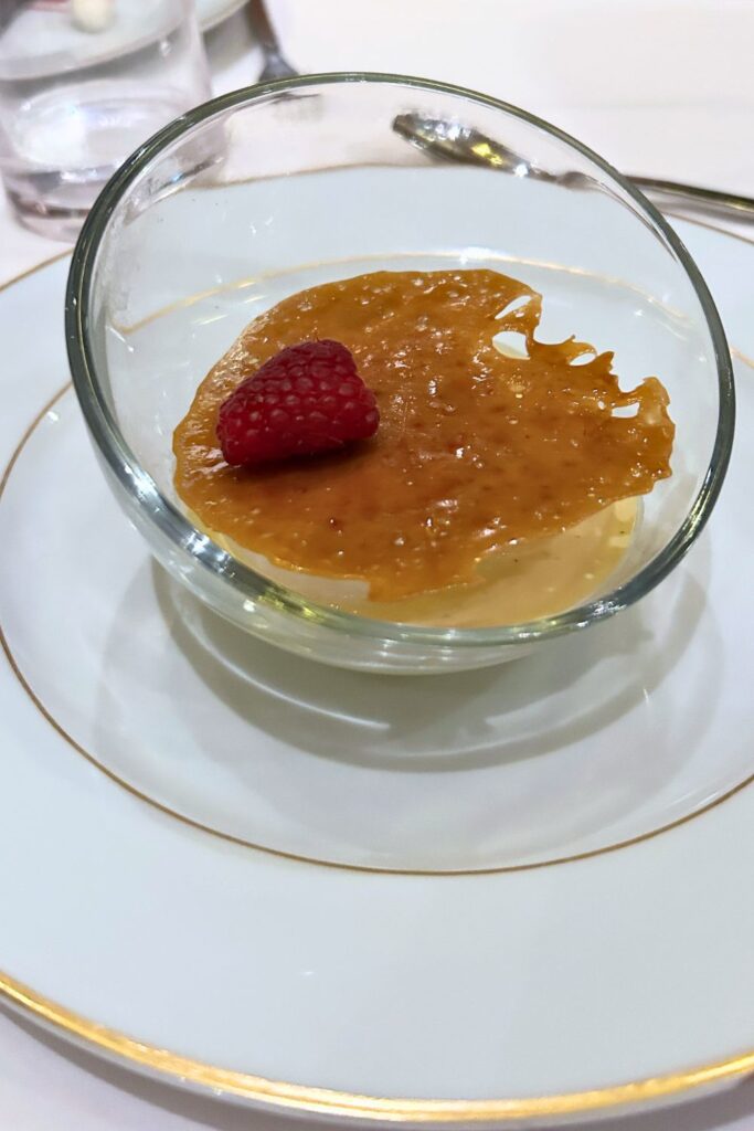 Creamy crème brûlée in a glass dessert bowl topped with a crisp caramelized sugar layer and fresh raspberry, served in the cruise ship dining room.