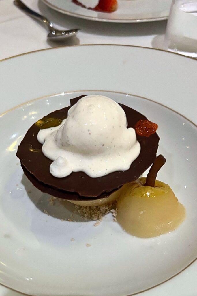 Warm chocolate dessert topped with melting vanilla ice cream and chocolate sauce, served with a poached pear on a cruise ship dinner plate.