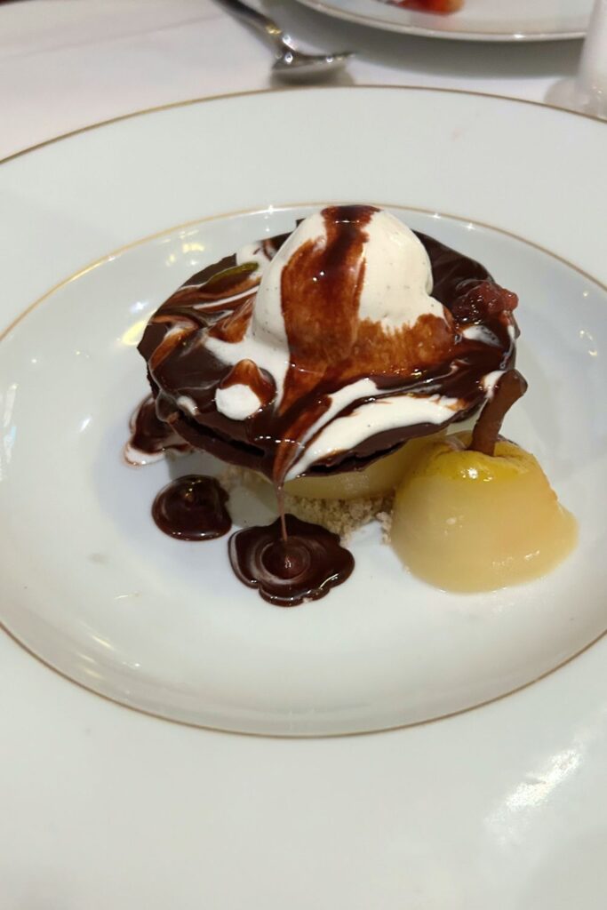 Chocolate dessert topped with vanilla ice cream and sauce, served with a poached pear on a cruise ship dining room plate.