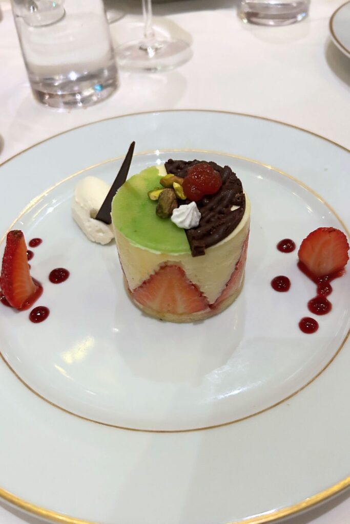 Elegant strawberry fraisier dessert with fresh strawberries, cream filling, pistachio topping, and chocolate garnish served in a cruise ship specialty restaurant.