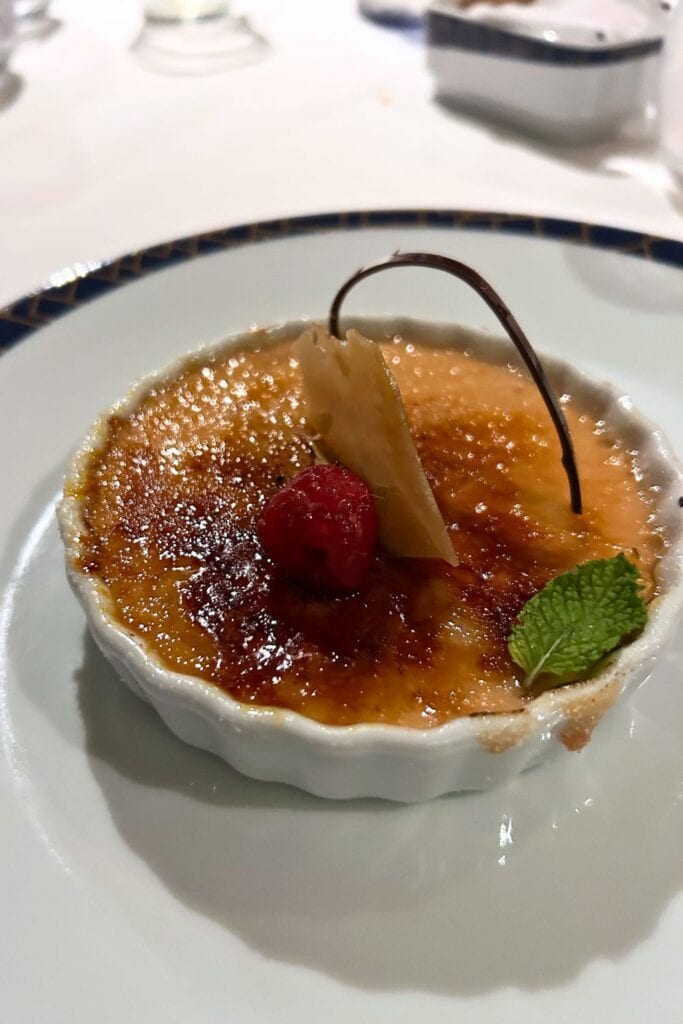 Crème brûlée dessert served on a cruise ship dining table with caramelized sugar topping, raspberry, mint leaf, and chocolate garnish.
